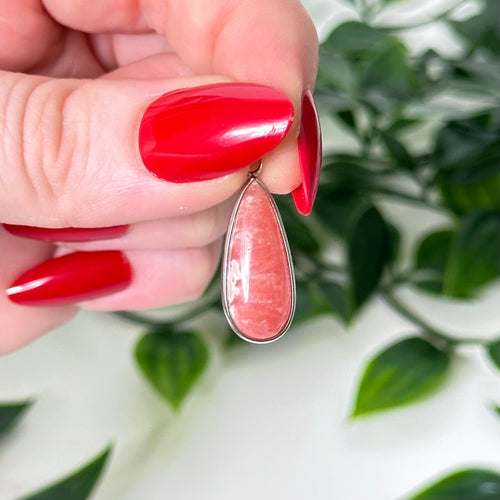 Pink crystal necklace pendant held by a hand with red nail polish against a green leafy background
