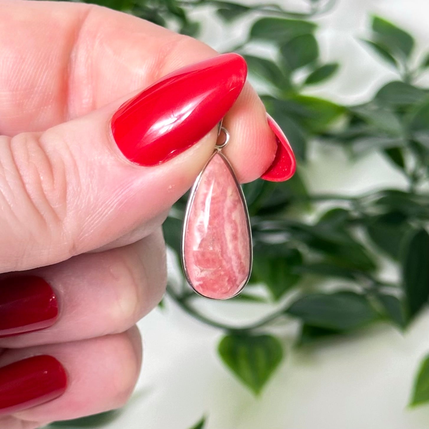 Rhodochrosite crystal pendant held by a hand against a green leafy background