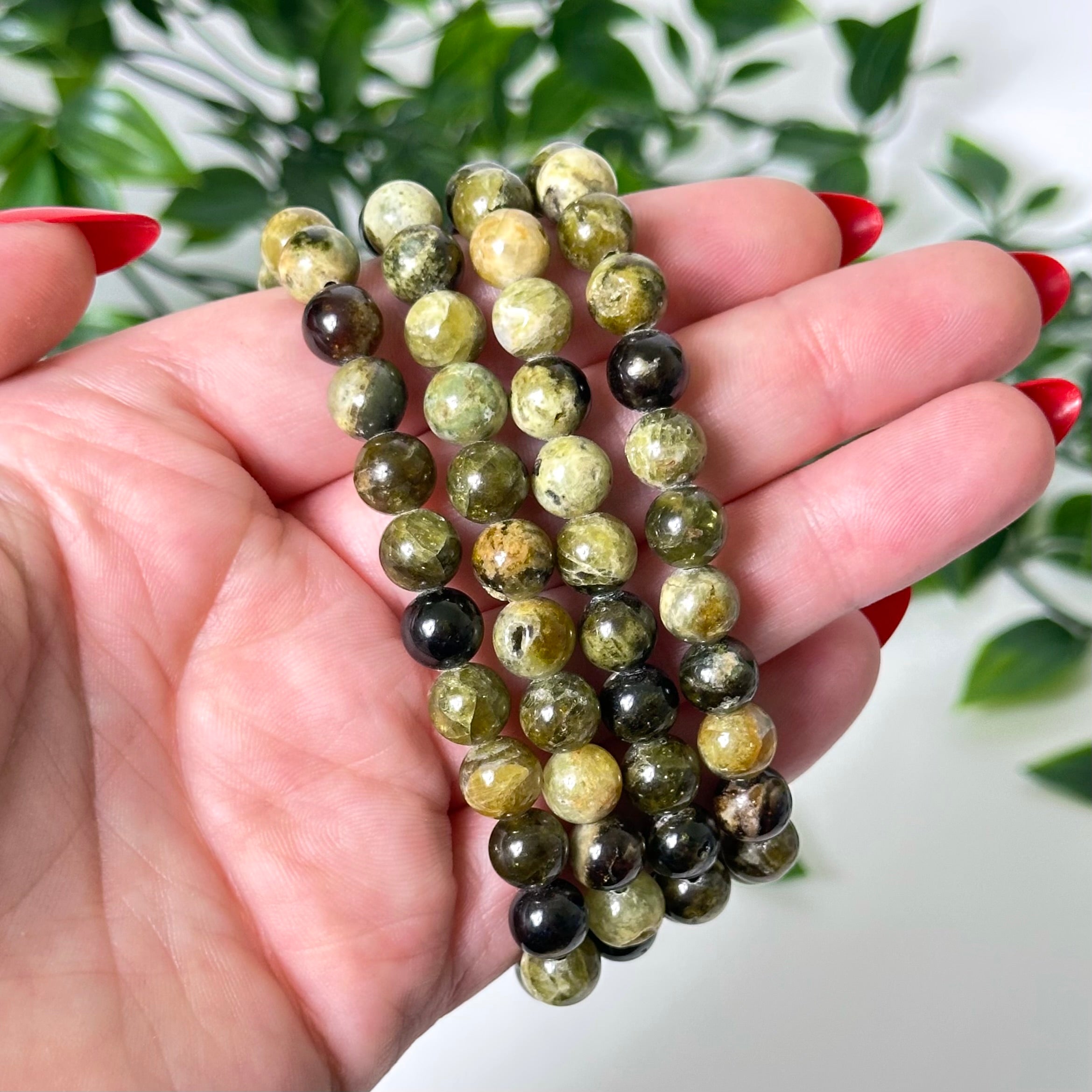 Hand holding a stack of green garnet crystal bead bracelets with a blurred green leafy background