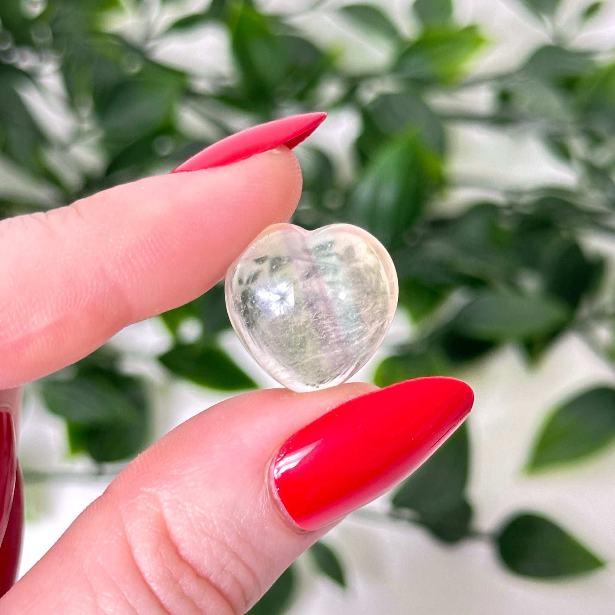 Heart-shaped crystal held between fingers with red nail polish against a green leafy background