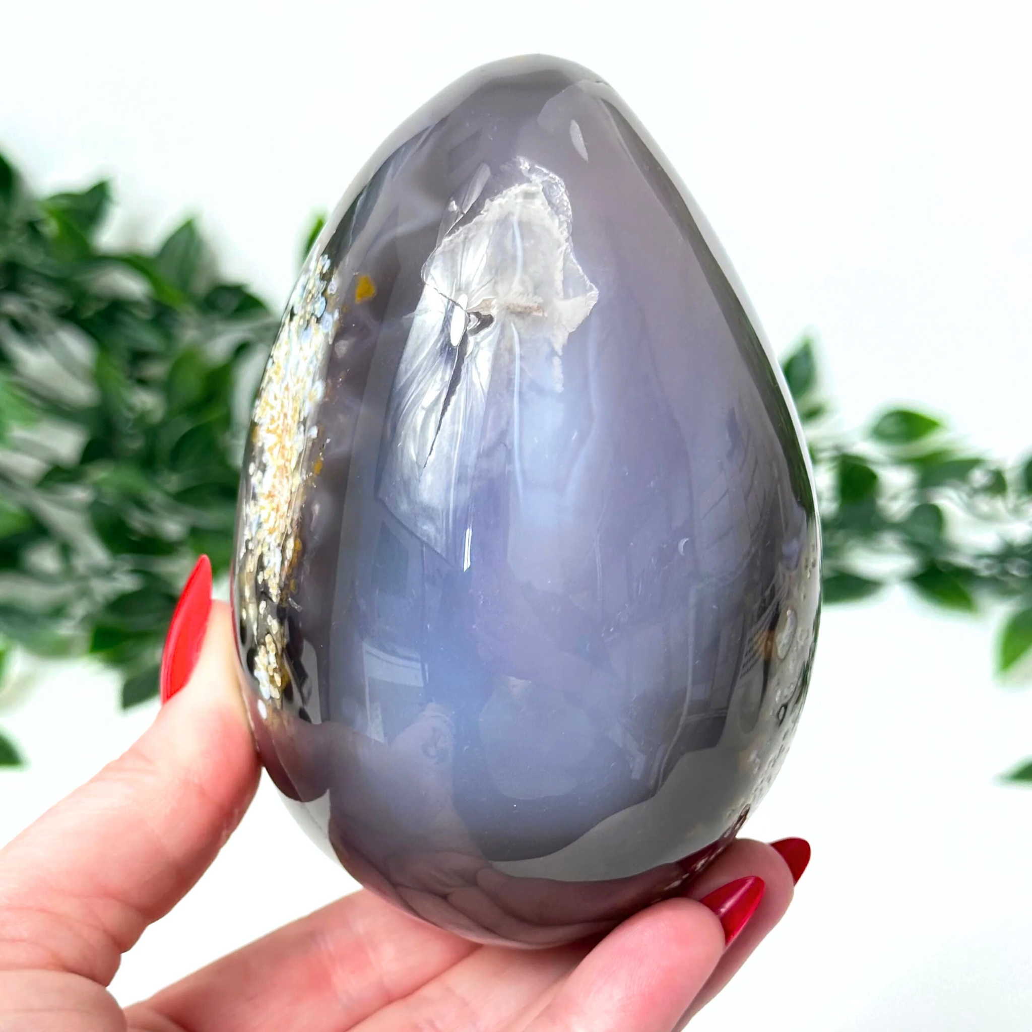 Hand holding a grey/blue agate crystal egg with a white center against a blurred green background
