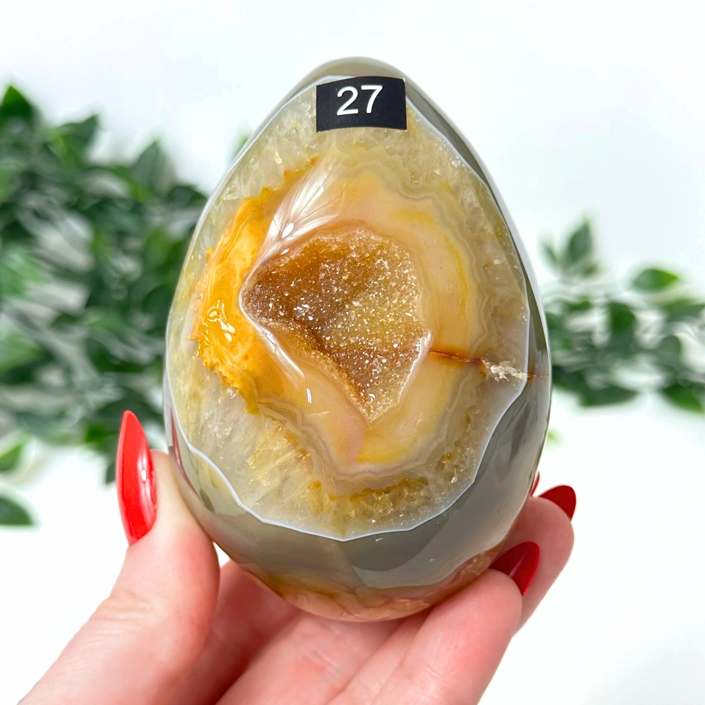 Hand holding a druzy agate egg with a yellow interior, against a blurred green and white background.