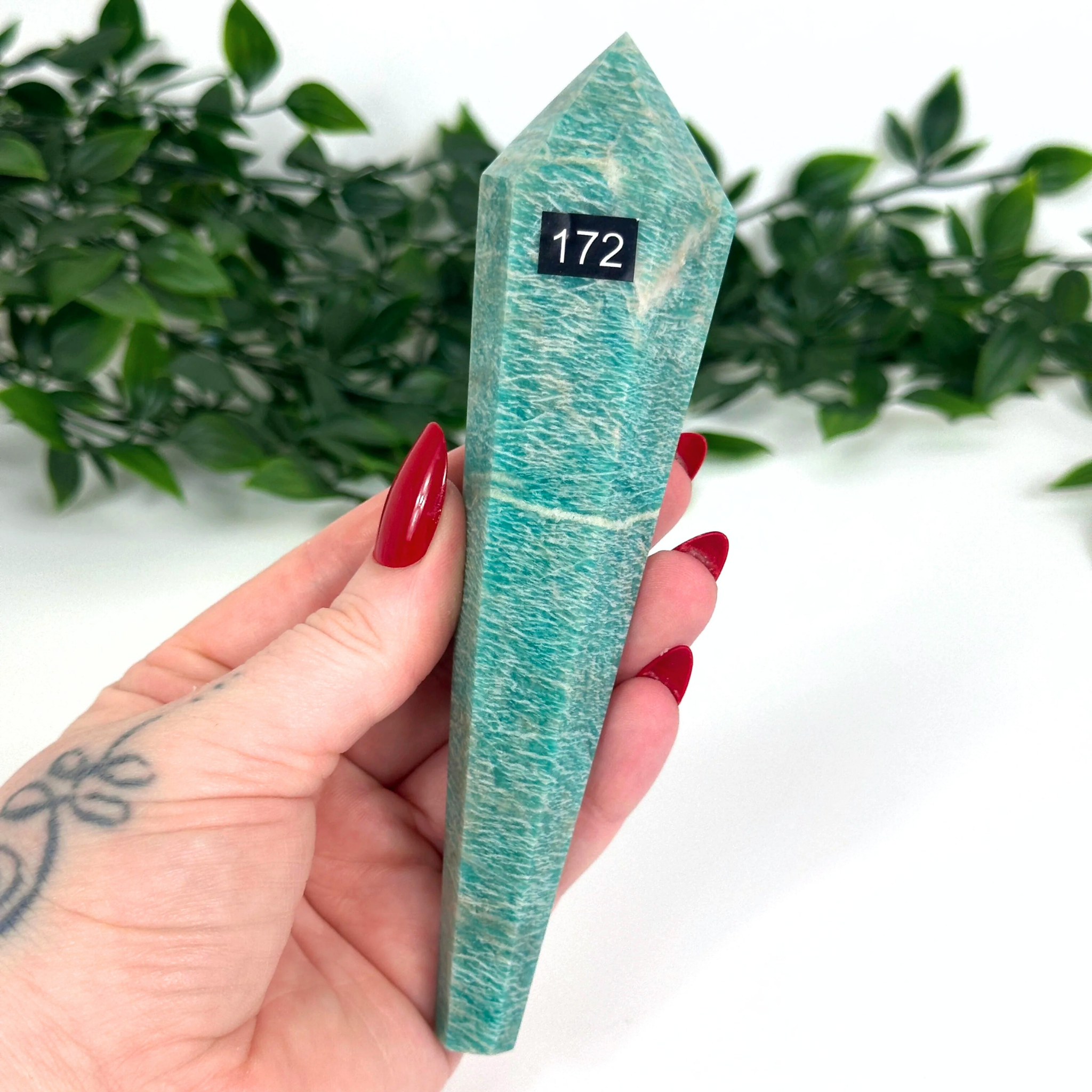 Amazonite crystal held by a hand with red nail polish against a white background with greenery.