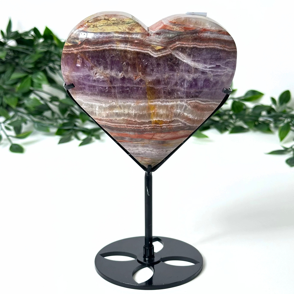 Heart-shaped multicolored amethyst stone on a black stand with a green leafy background