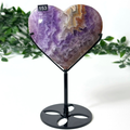 Heart-shaped purple amethyst stone on a black stand with greenery in the background
