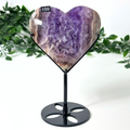 Heart-shaped amethyst crystal on a black stand with green foliage in the background