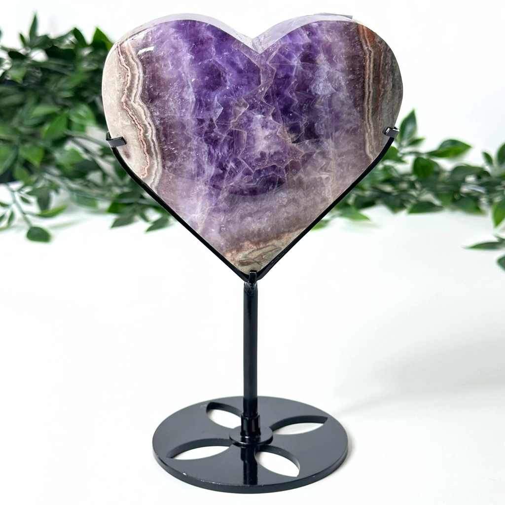 Heart-shaped amethyst crystal on a black stand with a green leafy background