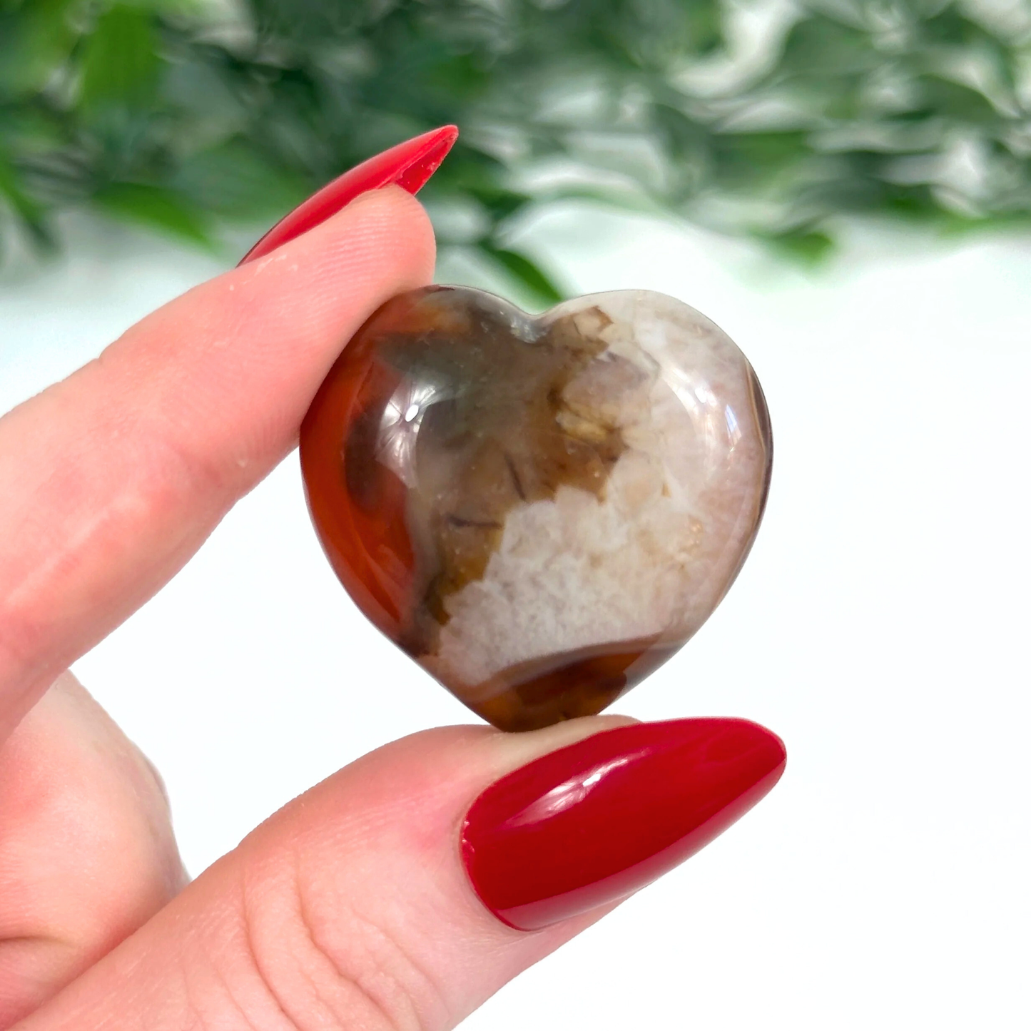 Heart-shaped carnelian crystal held between fingers with red nail polish against a blurred green background