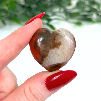 Heart-shaped carnelian crystal held between fingers with red nail polish against a blurred green background