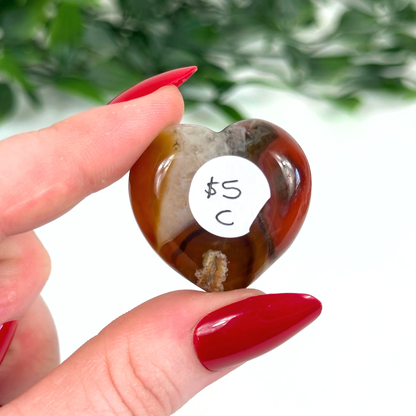 Heart-shaped carnelian crystal held by a hand with red nail polish, against a blurred green and white background.