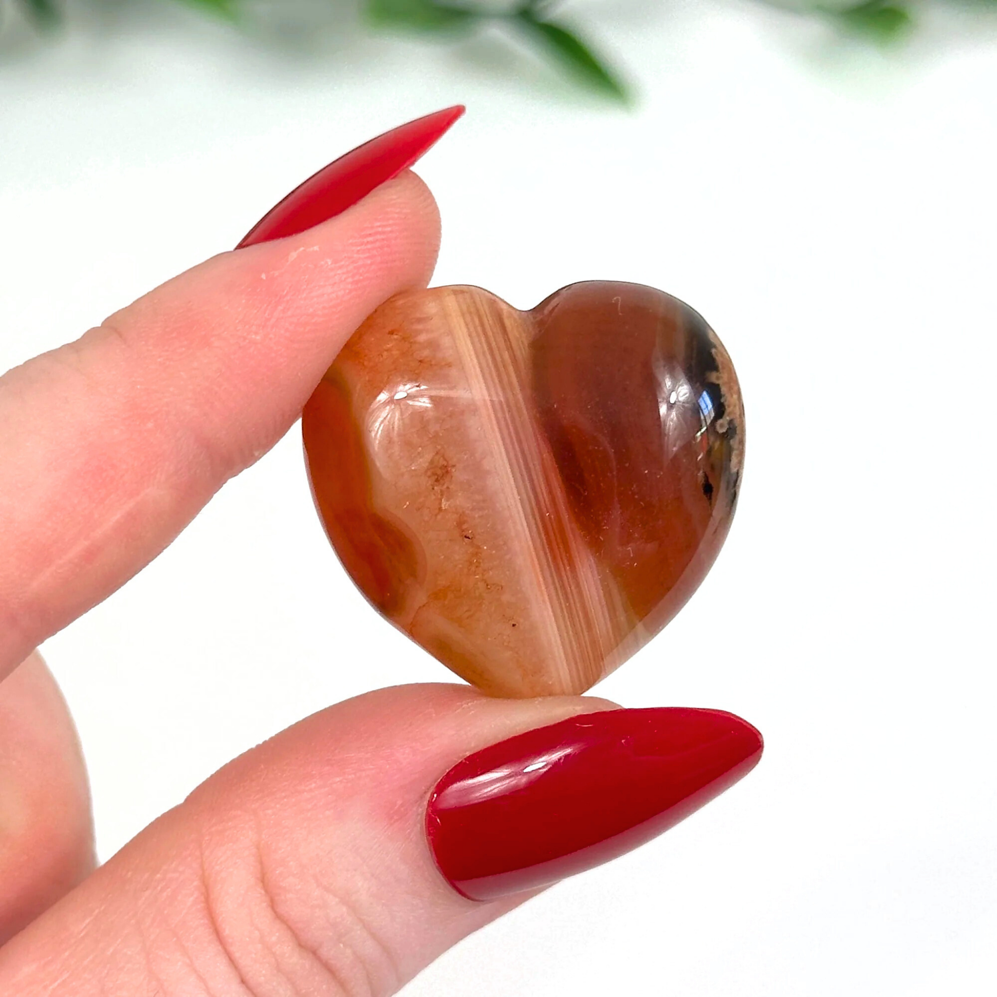 Heart-shaped carnelain crystal held between fingers with red nail polish on a white background