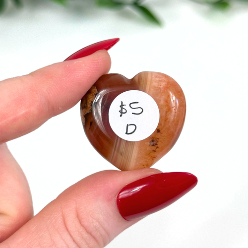 Heart-shaped carnelian crystal held by a hand with red nail polish.