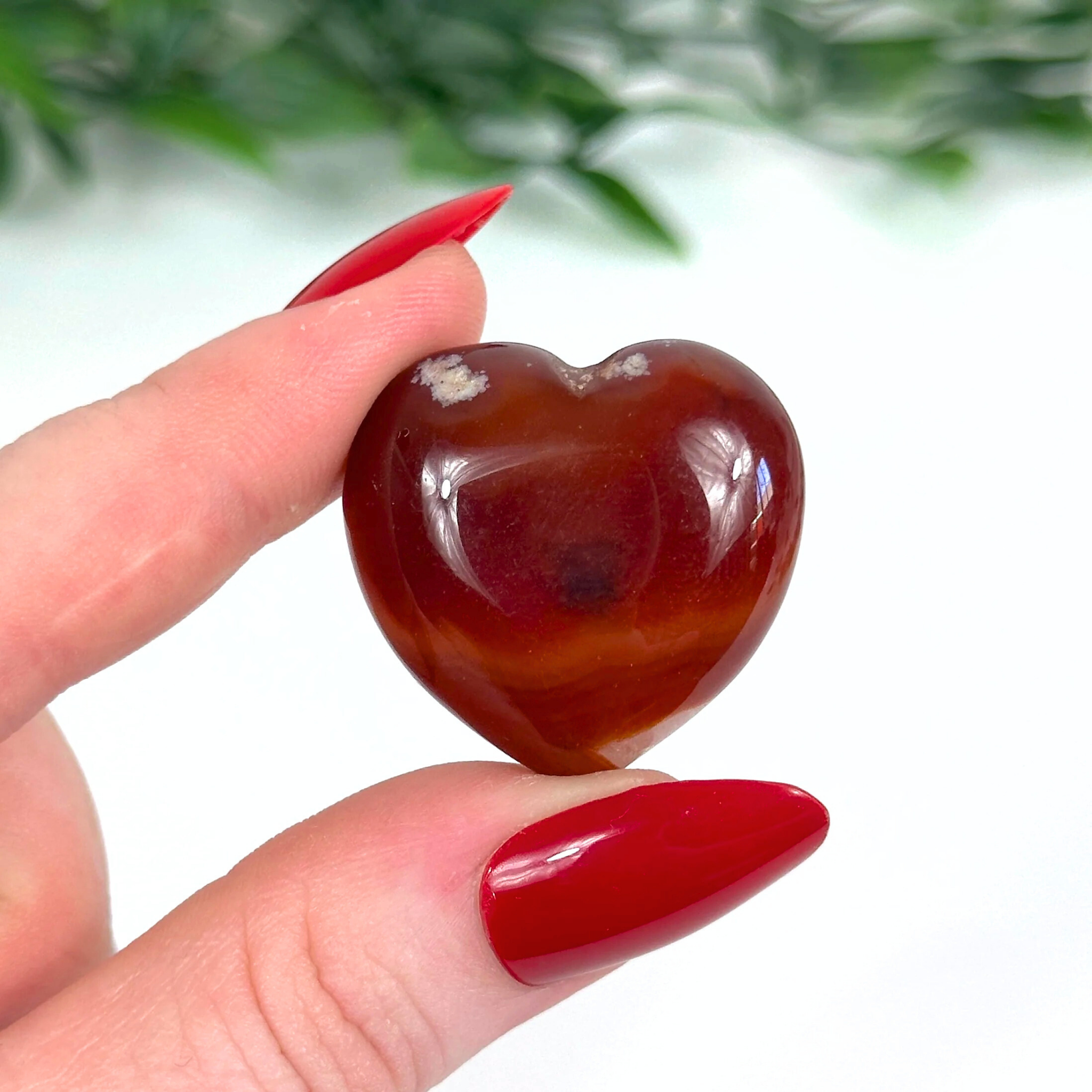 Heart-shaped carnelian crystal held by a hand with red nail polish against a blurred green and white background