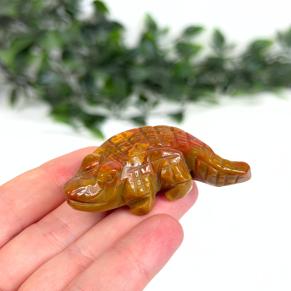 Hand holding a mookaite crocodile carving with a blurred green foliage background