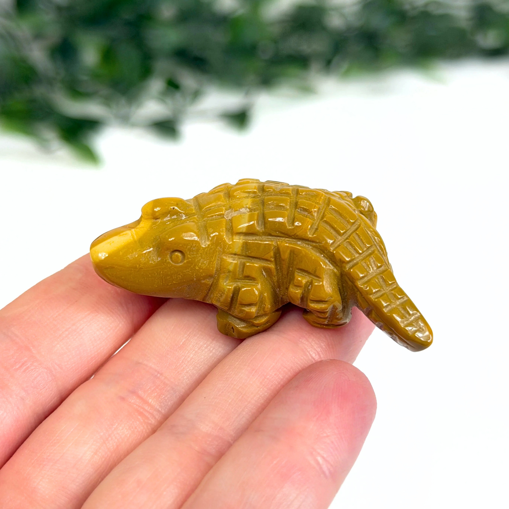 Hand holding a mookaite crocodile carving with a blurred green and white background