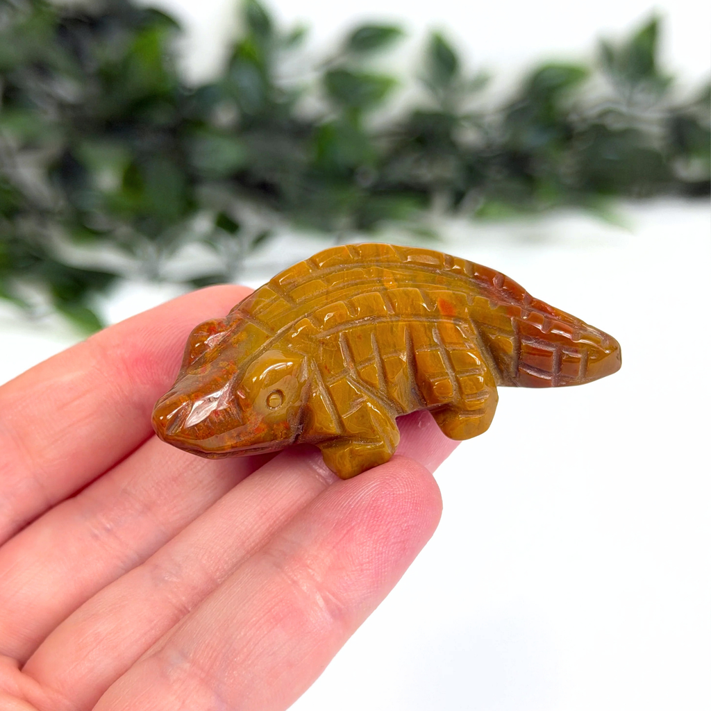 Mookaite crocodile carving held in a hand with a blurred green and white background