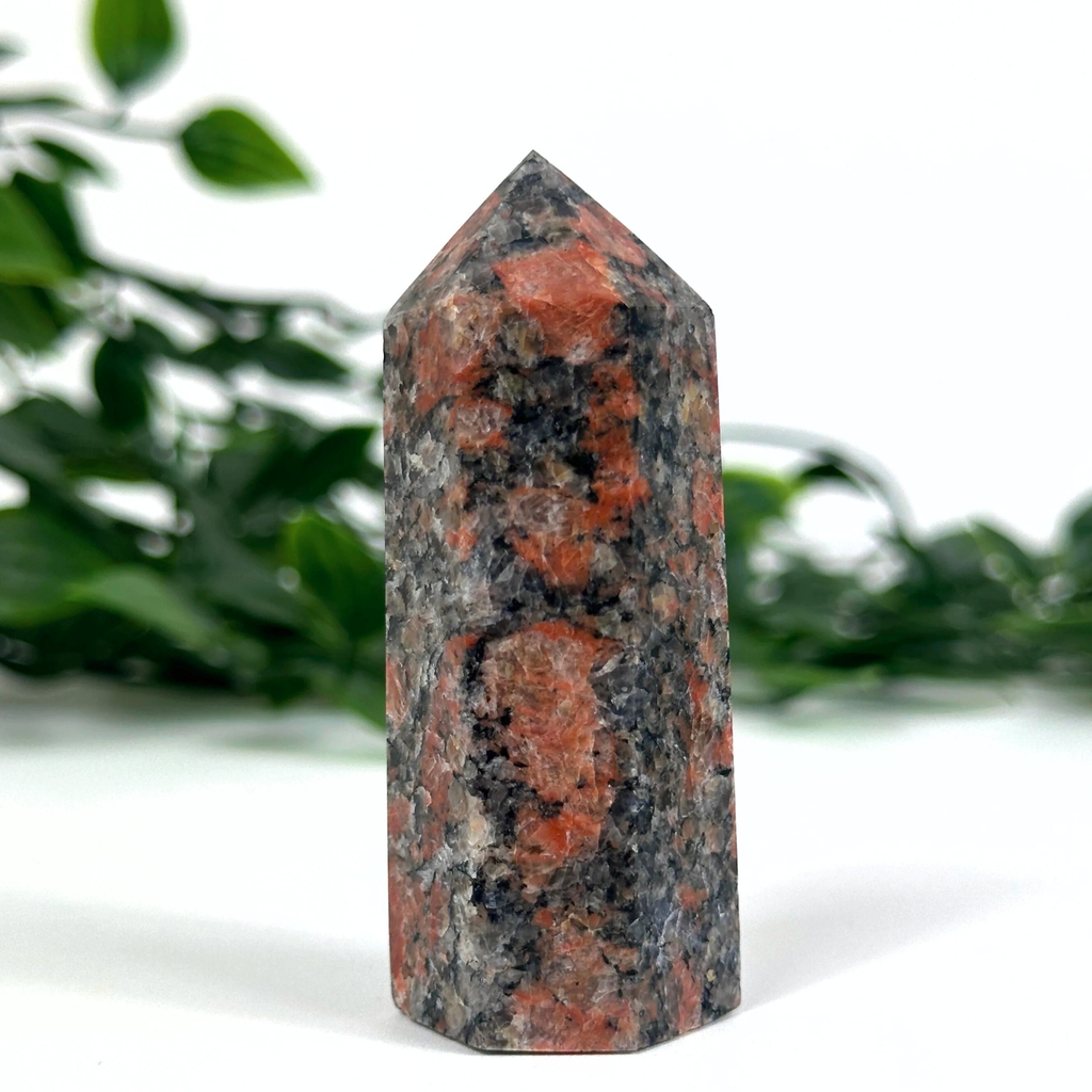 Feldspar crystal tower with red and black patterns on a white surface with green foliage in the background