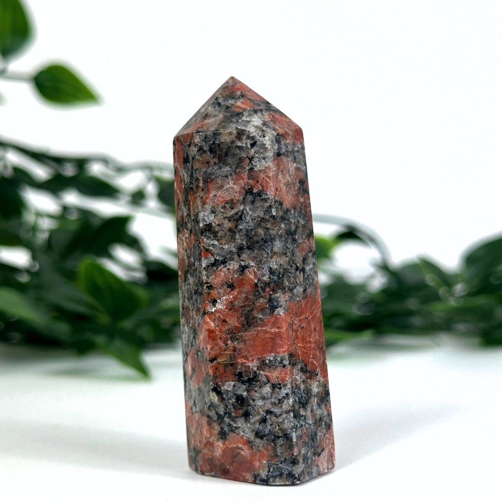 Feldspar crystal tower with red and gray patterns on a white surface with green leaves in the background