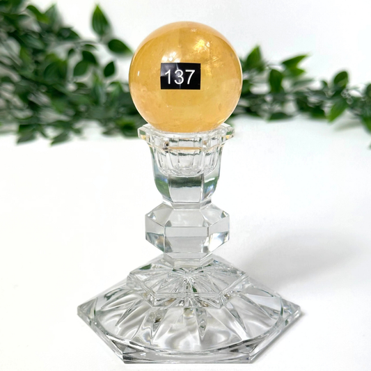 Crystal pedestal with a honey calcite sphere on top, against a white background with green leaves.