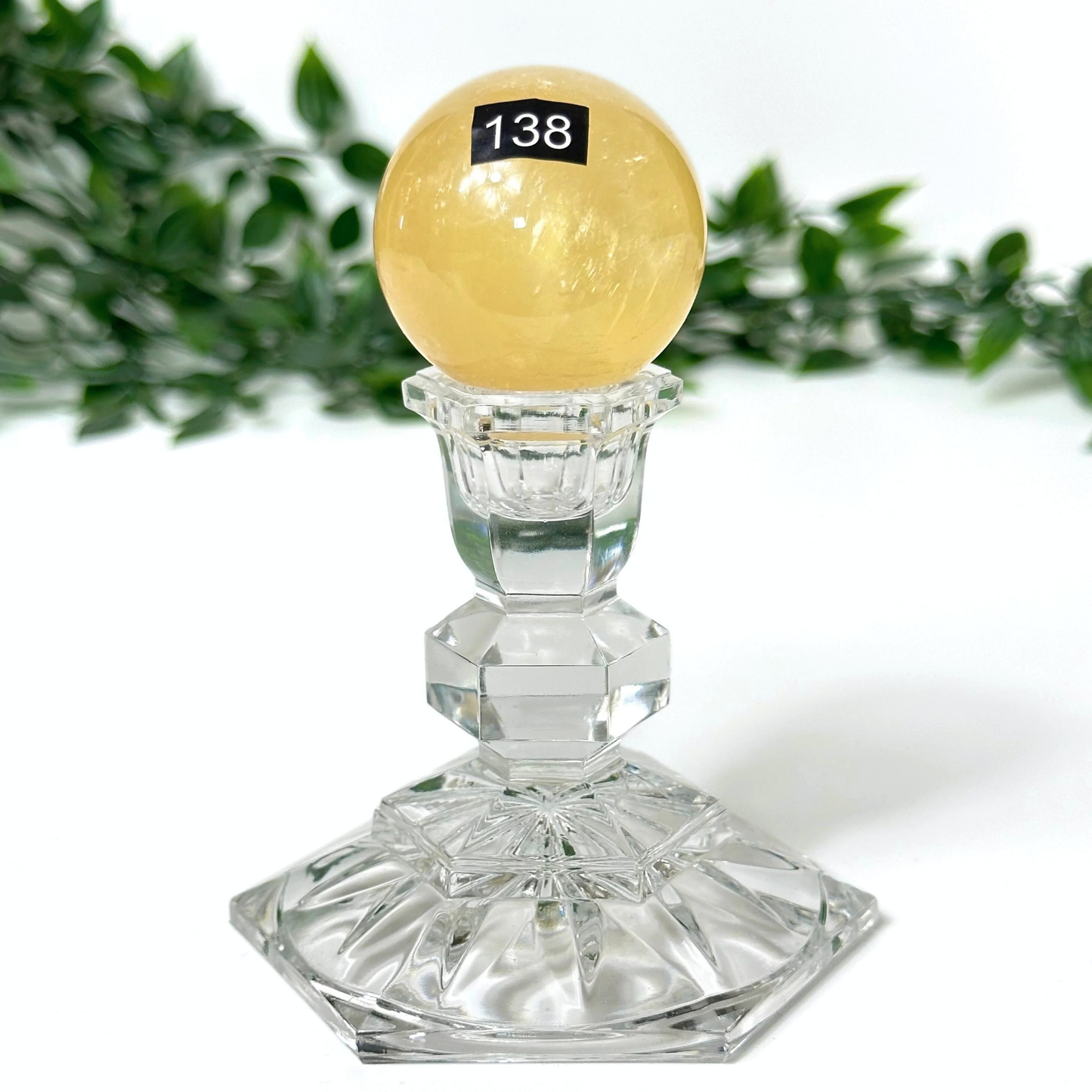 Honey calcite crystal sphere on a crystal stand with green foliage in the background