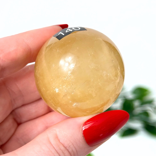 Hand holding a honey calcite sphere against a blurred green plant background