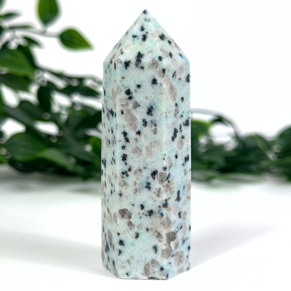 Speckled kiwi jasper crystal tower on a white surface with greenery in the background