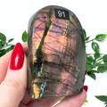 Person holding a labradorite crystal with colorful patterns, surrounded by green leaves.