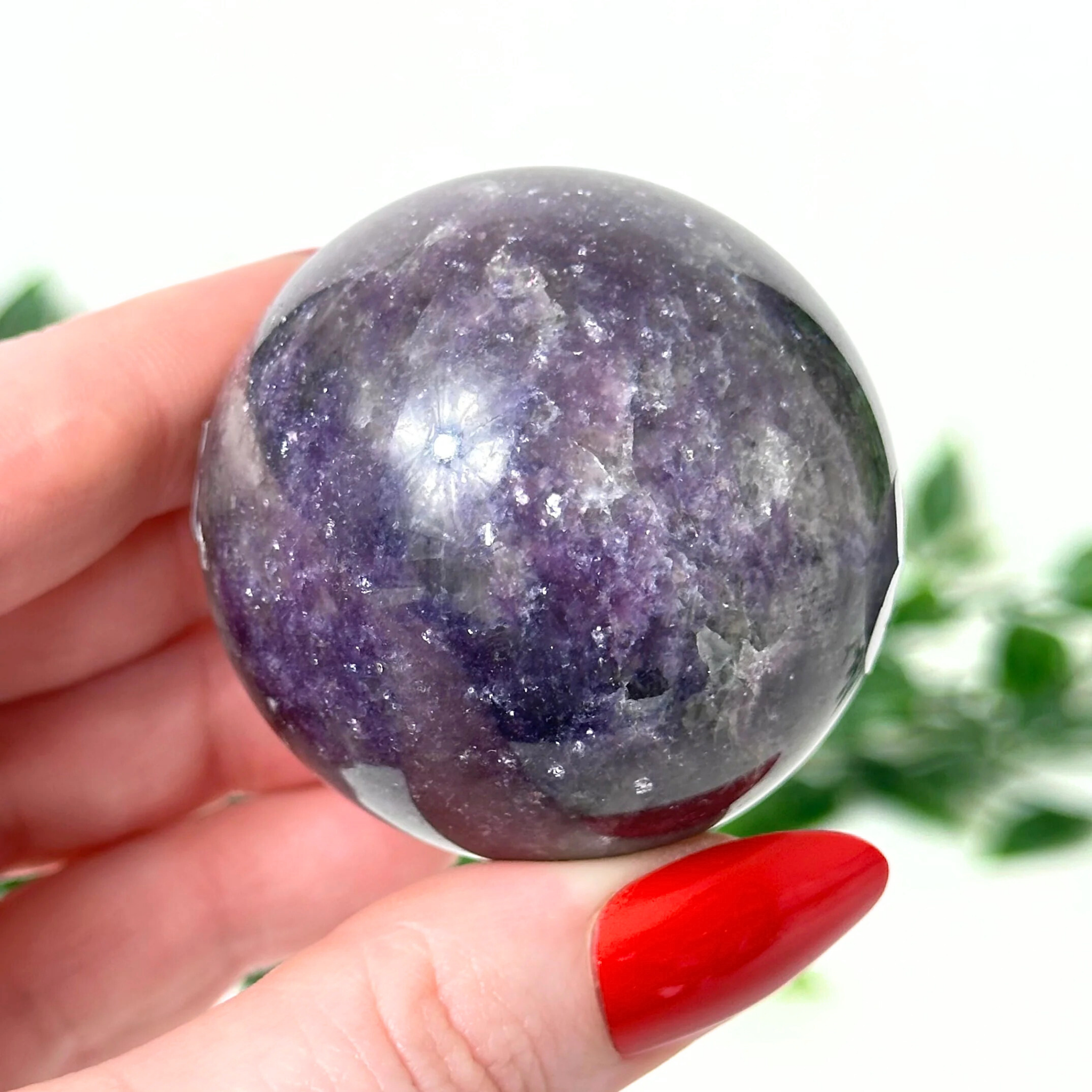 Hand holding a purple and gray lepidolite crystal ball with a blurred green background