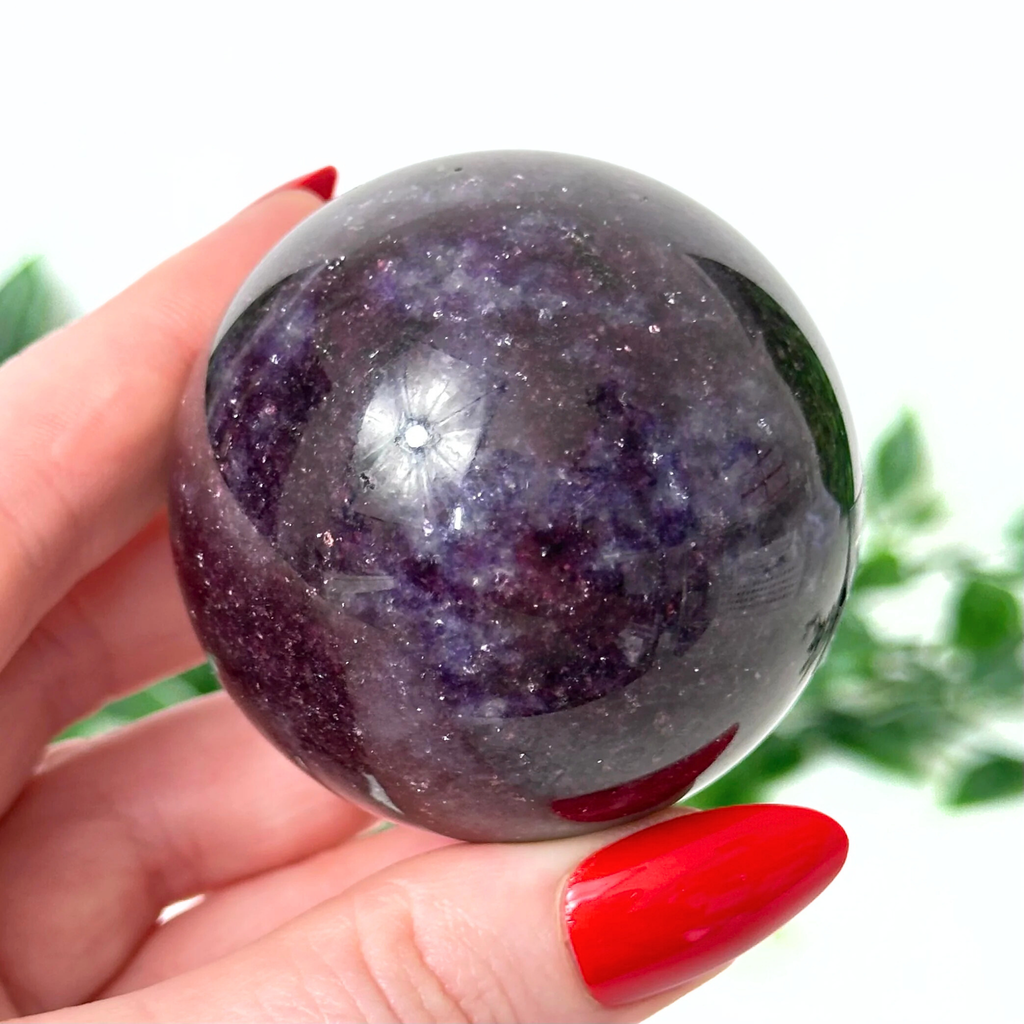 Hand holding a purple lepidolite crystal ball with a blurred green plant background