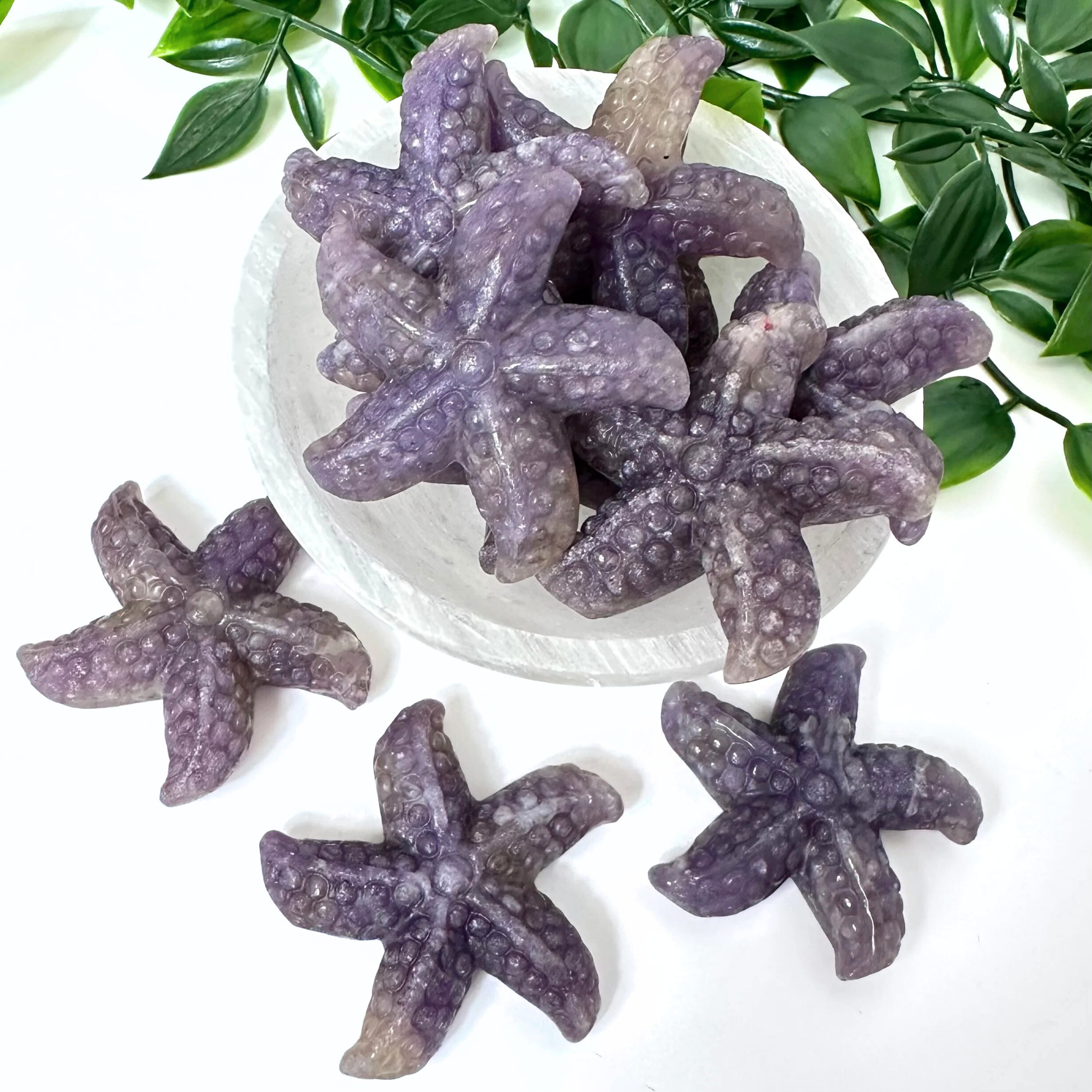 Lepidolite starfish crystal carvings on a white plate with green leaves in the background