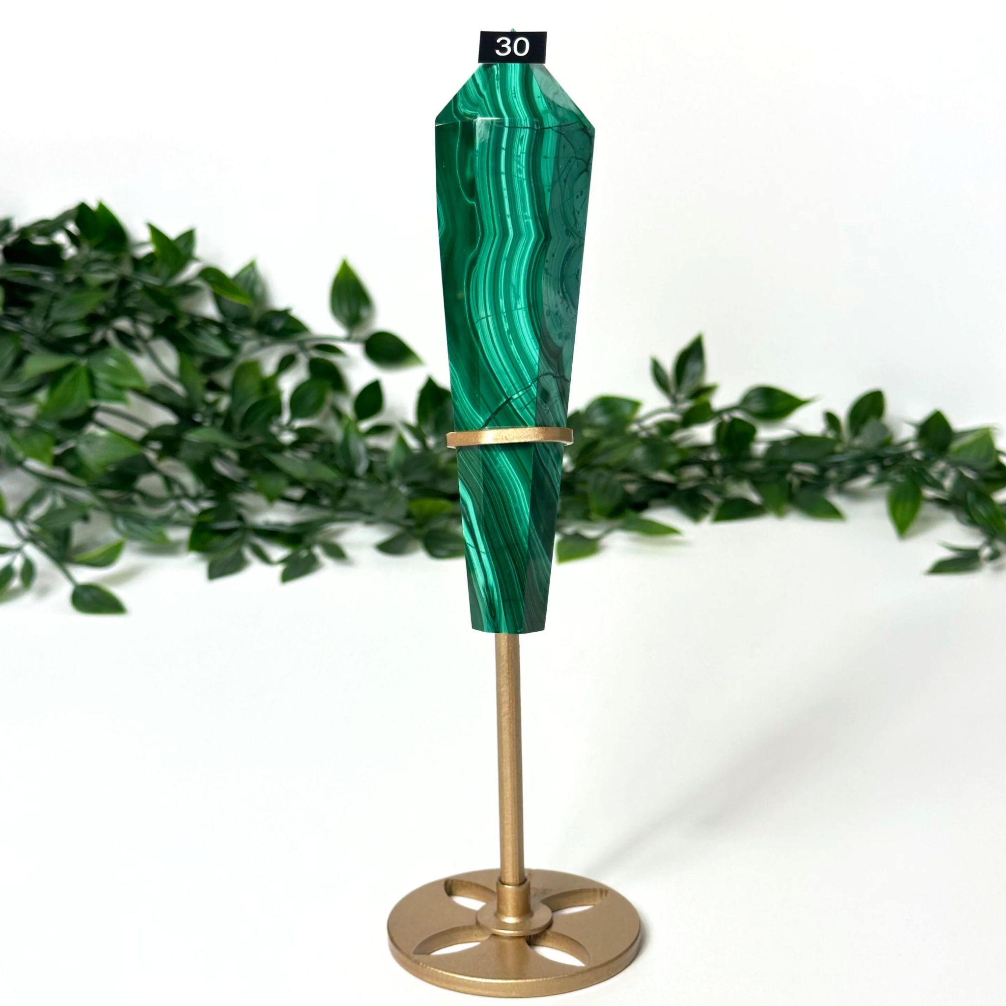 Green malachite crystal wand with gold stand on a white and blurred leaf background