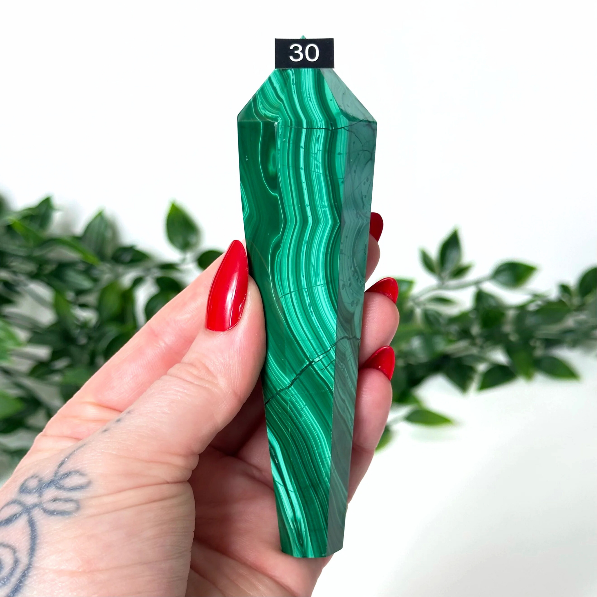 Hand holding a green malachite crystal wand on a white and blurred leaf background