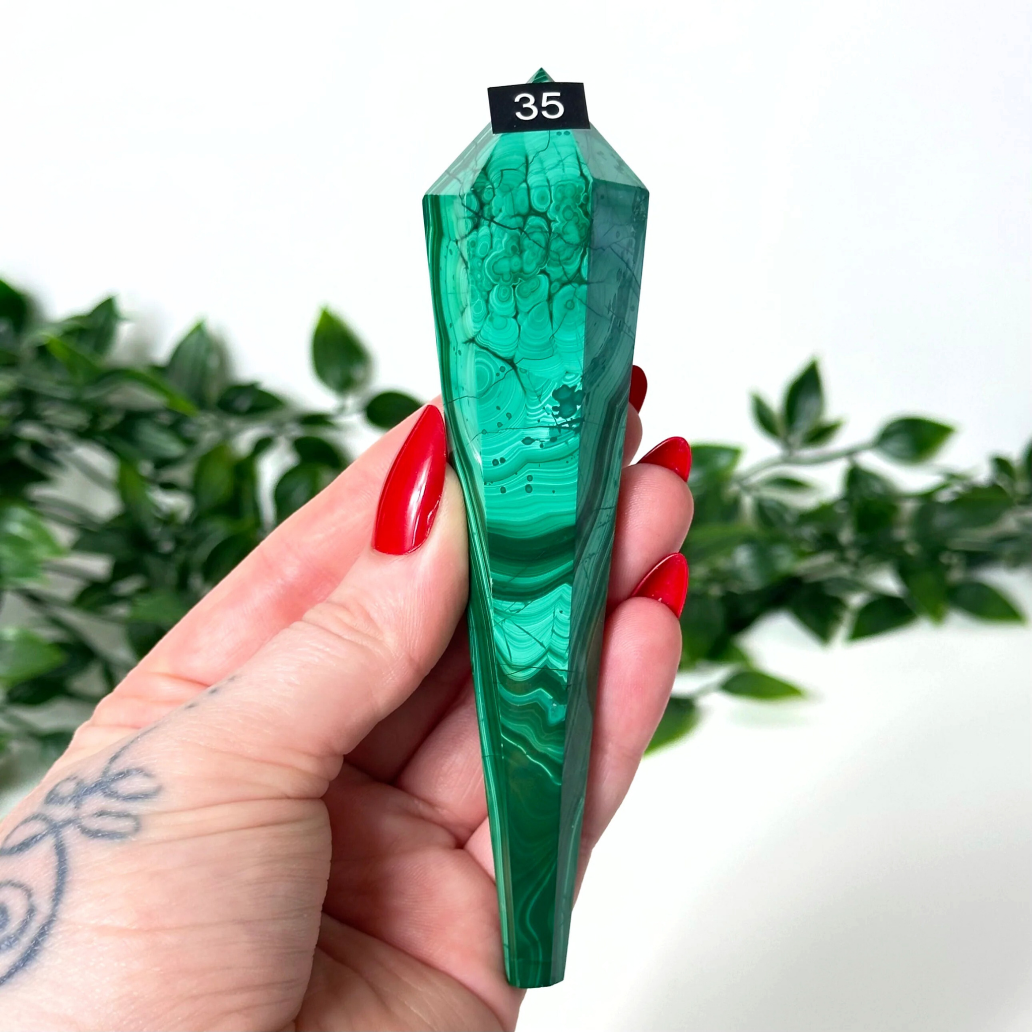 Green malachite crystal held by a hand with red nails against a white background with green leaves.