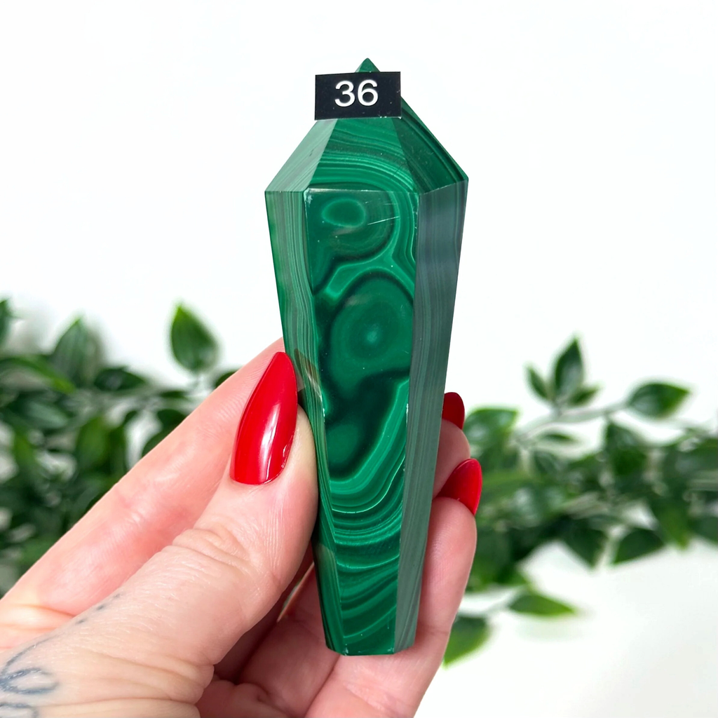 Green malachite crystal held by a hand with red nails against a blurred green plant background