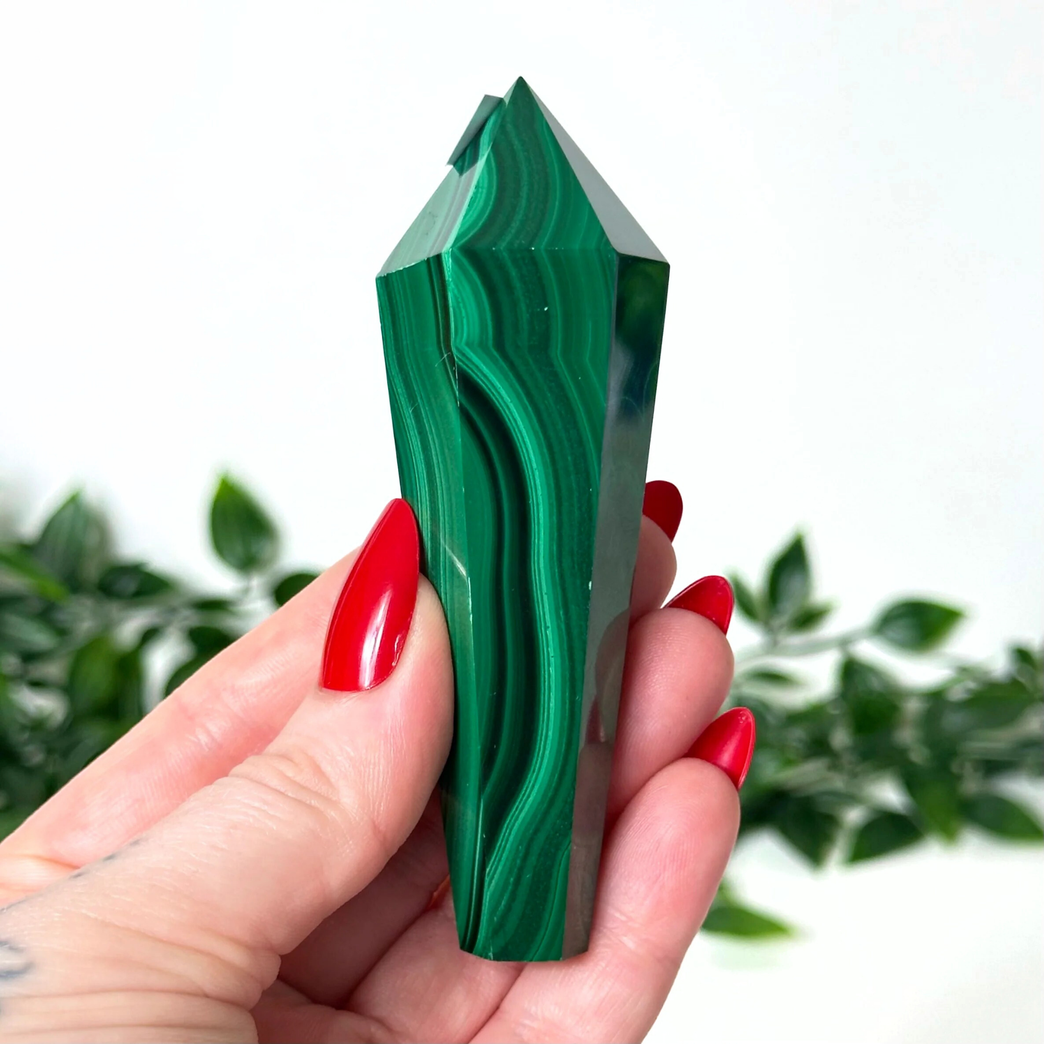 Green malachite crystal held in a hand with red nail polish against a blurred green leafy background