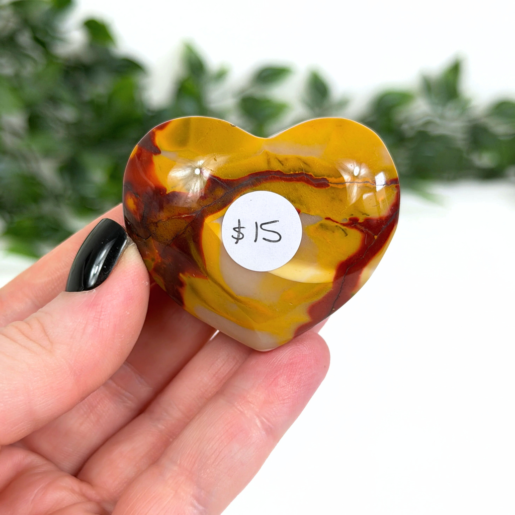 Heart-shaped mookaite stone held in a hand with a $15 label, blurred greenery in the background