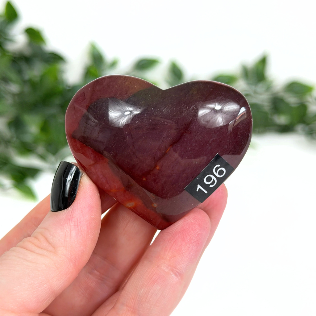 Heart-shaped red mookaite stone held in a hand with a blurred green leafy background