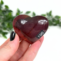 Heart-shaped red mookaite stone held in a hand with a blurred green leafy background
