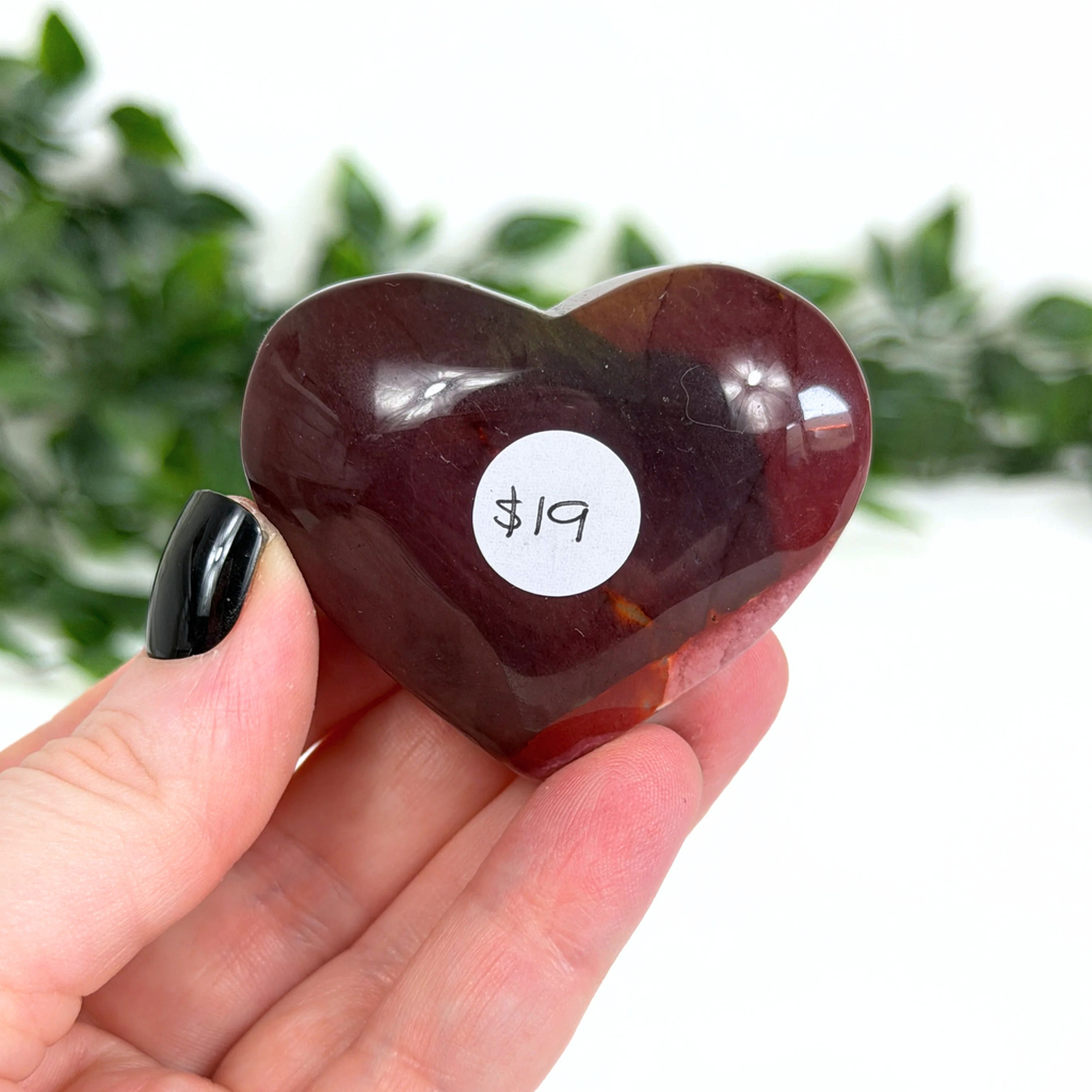 Heart-shaped dark red mookaite stone held in a hand with a price tag sticker, against a blurred green leafy background.