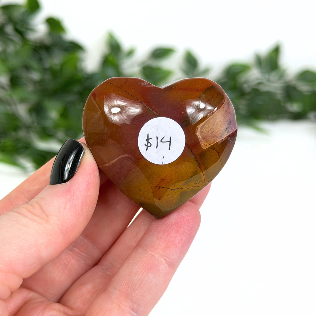 Heart-shaped brown mookaite crystal held in a hand with a price tag label, against a blurred green foliage background.