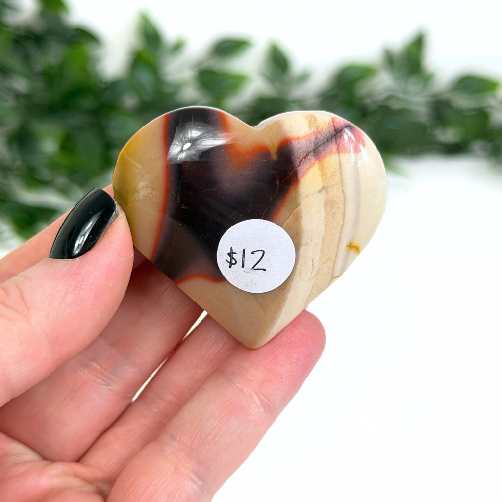 Heart-shaped mookaite stone with a price tag held in a hand against a blurred green background