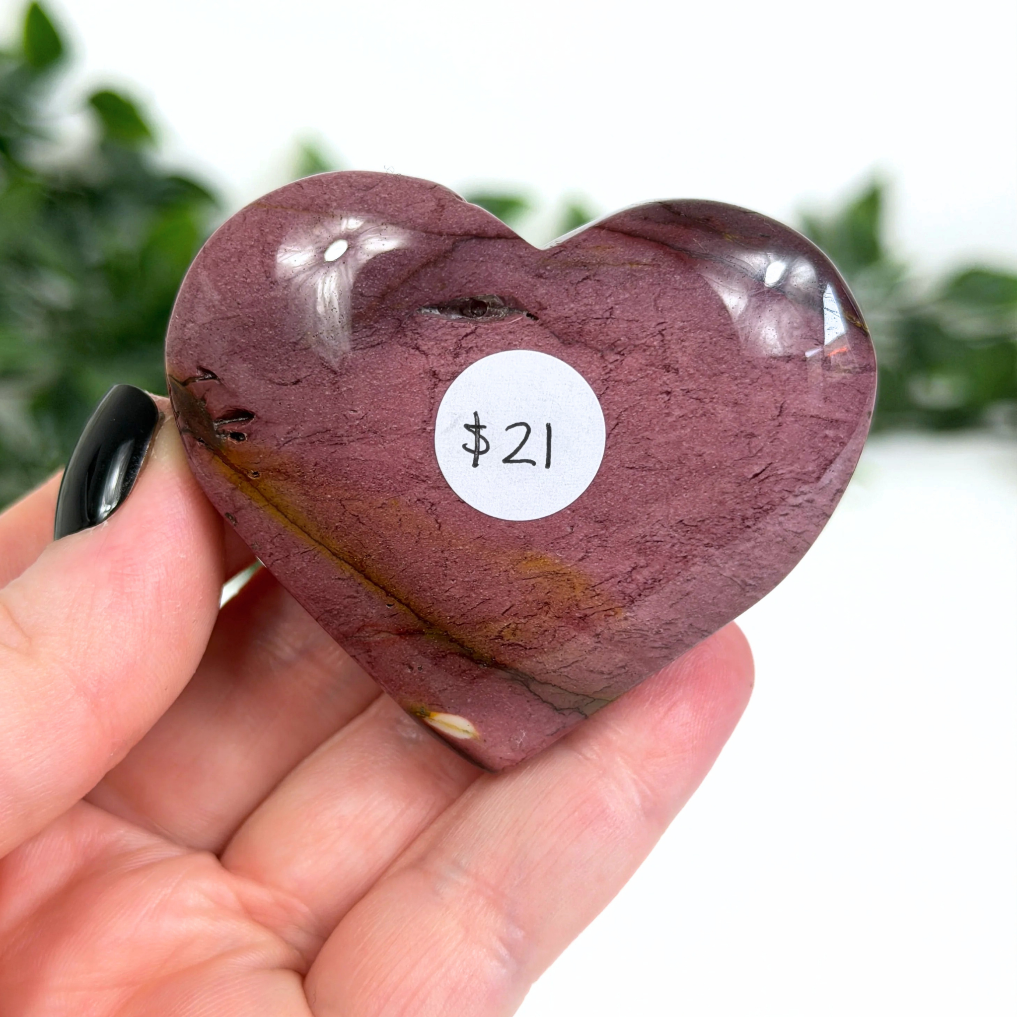 Heart-shaped purple mookaite stone held in a hand with a price tag of $21 on a blurred green background