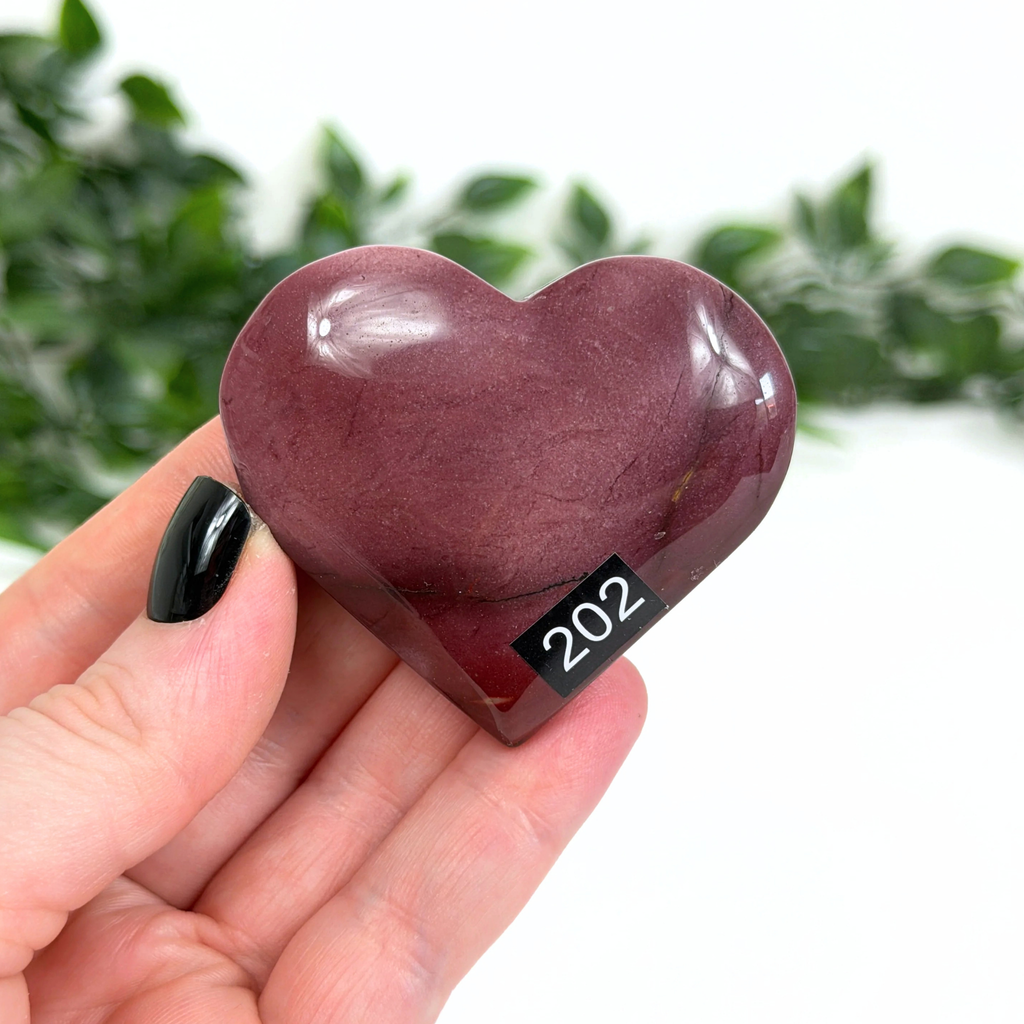 Heart-shaped purple mookaite stone held in a hand with a blurred green leafy background