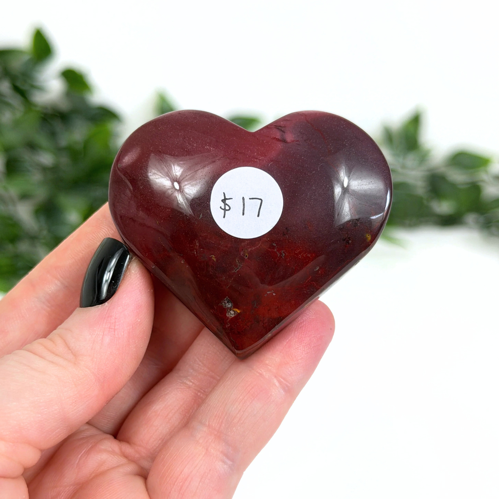 Heart-shaped mookaite crystal held in a hand with a white label displaying "$17" against a blurred green background.