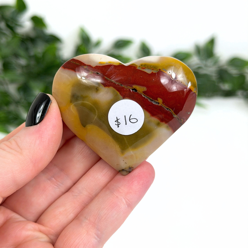 Heart-shaped marbled mookaite stone held in a hand with a price tag of $16, against a blurred greenery background.