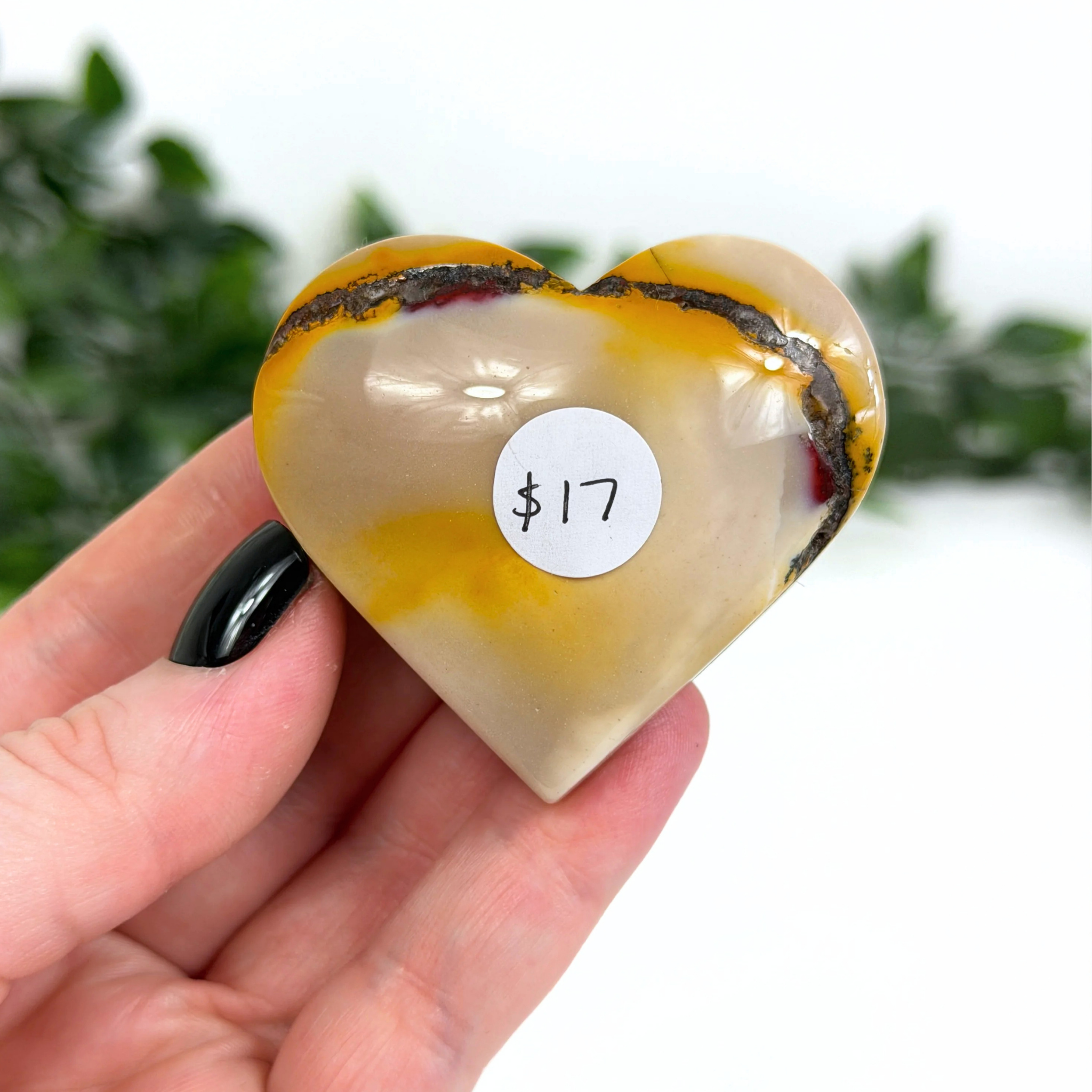 Heart-shaped mookaite crystal with a price tag held by a hand against a blurred green background