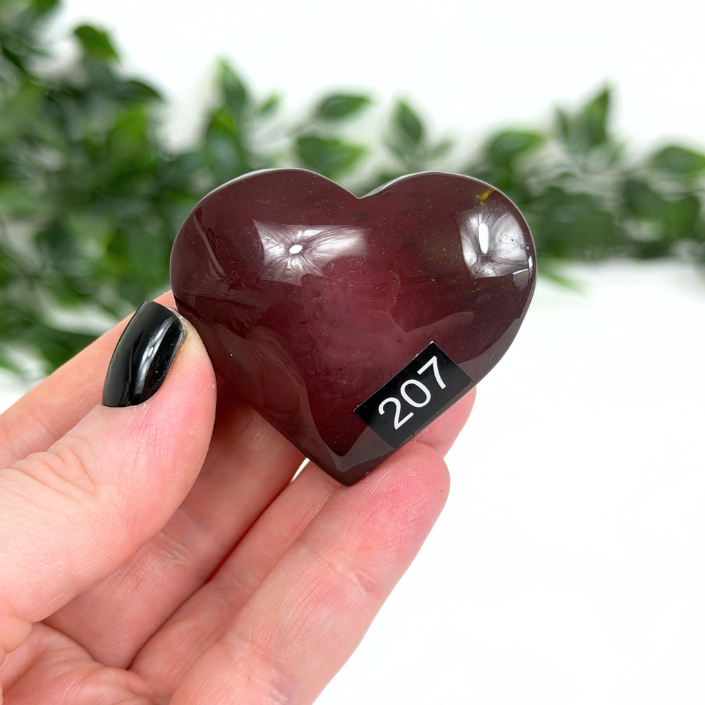 Heart-shaped dark red mookaite stone held in a hand with a blurred green background