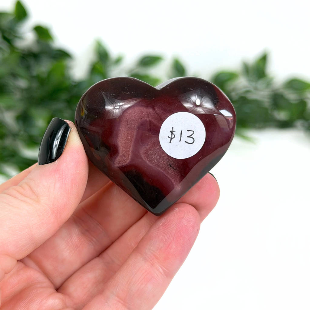 Heart-shaped dark red mookaite stone held in a hand with a white label indicating $13, against a blurred green leafy background.