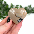 Heart-shaped brown mookaite crystal held by a hand with black nail polish, against a blurred green and white background.