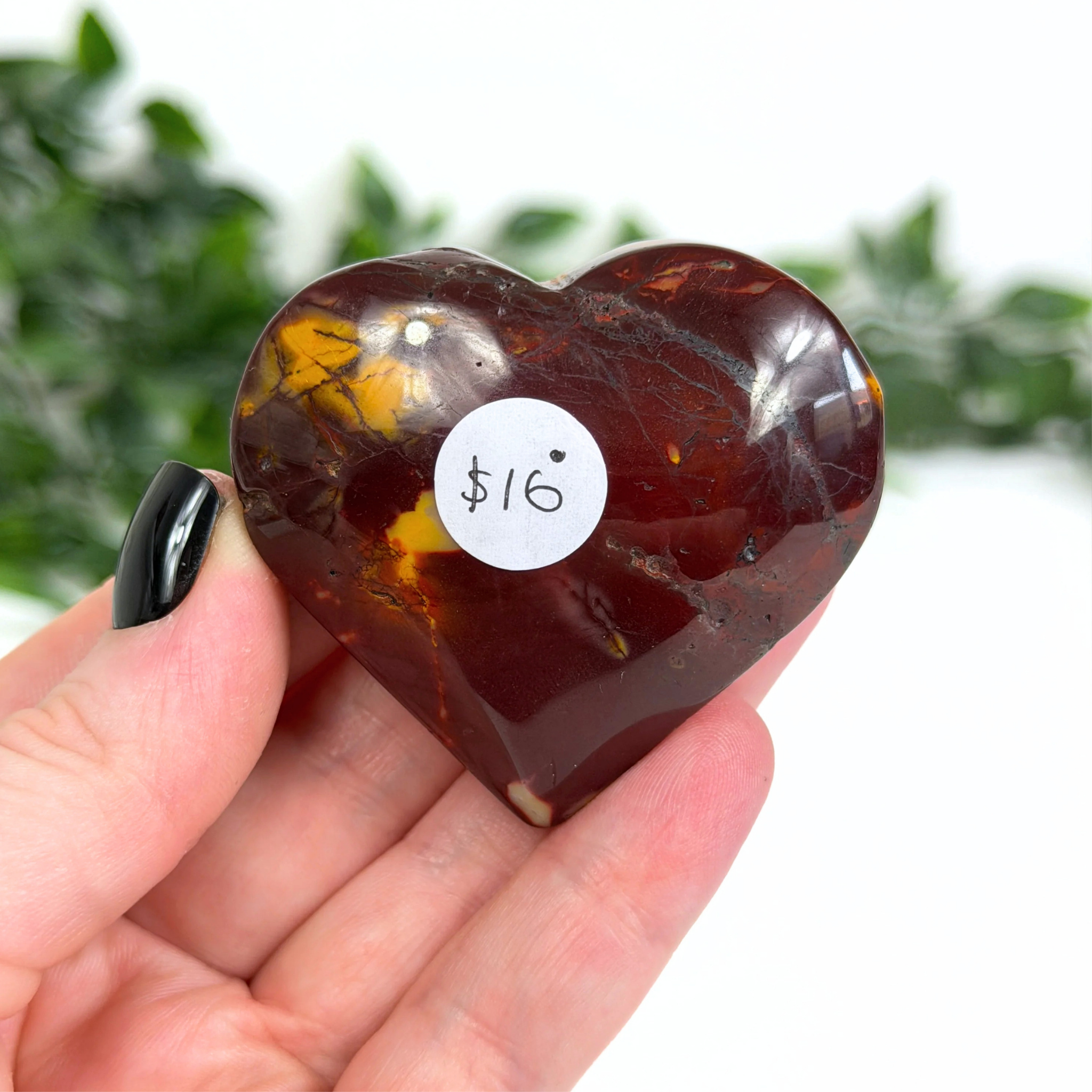 Heart-shaped mookaite stone held in a hand with a price tag of $16, against a blurred green background.